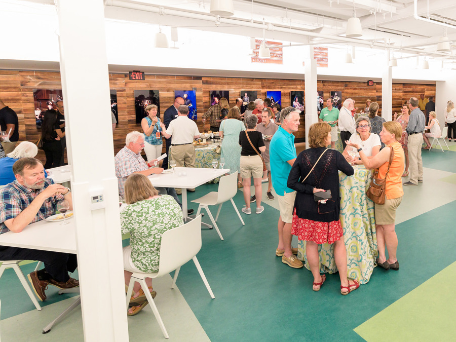 Large group of guests mingling in a bright reception space with tables and wood-paneled walls displaying performance photos.