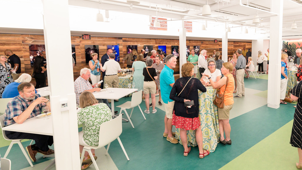 Large group of guests mingling in a bright reception space with tables and wood-paneled walls displaying performance photos.