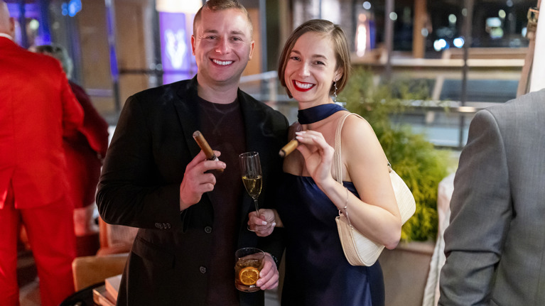 Two guests holding cigars and drinks at an outdoor lounge area.