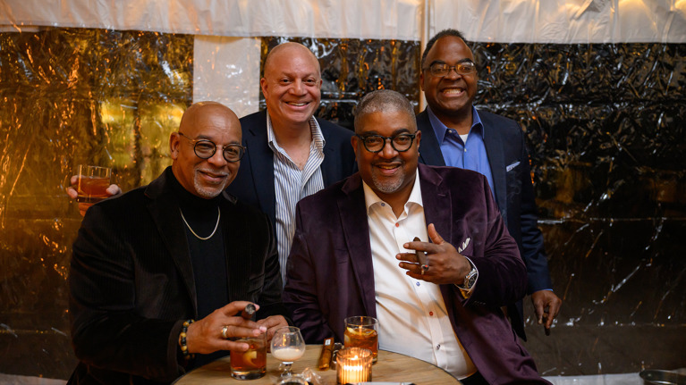 Group photo of four people standing outdoors at night in front of festive lights, one smoking a cigar.