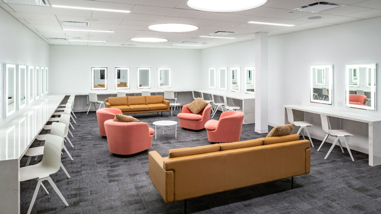 Spacious dressing room with lighted mirrors, white counters, and seating areas with pink and tan sofas.