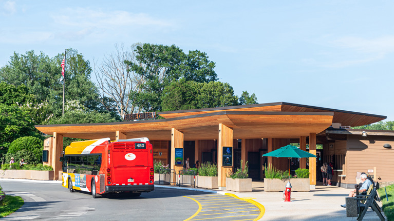 Shuttle bus arriving at Filene Center main gate with wooden canopy and landscaped entrance.