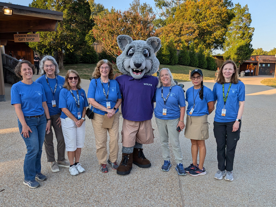 A group of volunteers wearing blue shirts pose with Wolf Trap's mascot Wolfie on the plaza, with the gift shop and greenery in the background.