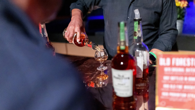 Close-up of bourbon being poured into a glass on a bar with several bottles in view.