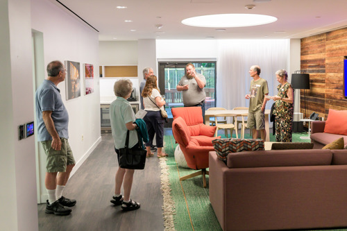 Group of people touring a renovated artist lounge featuring modern furniture, wood accent wall, and bright lighting.