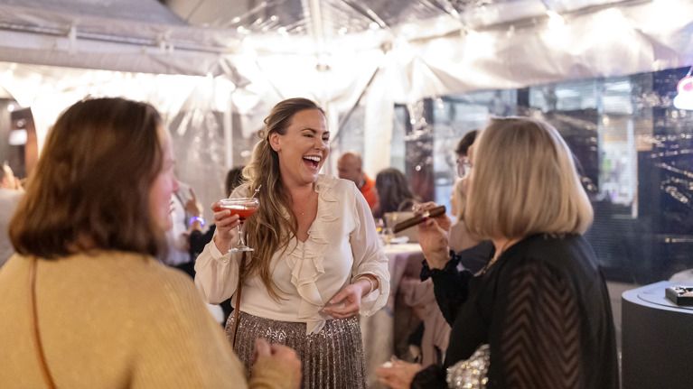 Guests chatting under a clear tent with string lights, holding cocktails during the Bourbon and Bubbles event.