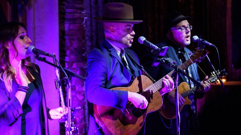 Three musicians singing and playing guitar with microphones.
