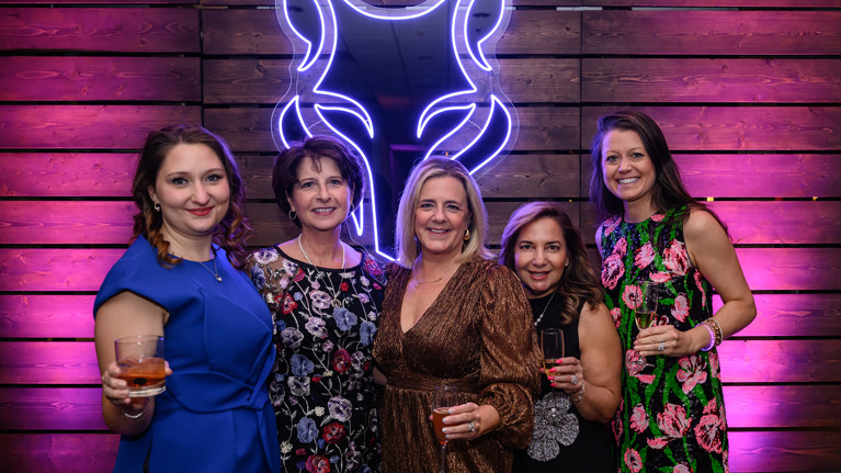 Group of guests posing in front of a neon wolf logo on a wooden backdrop, holding drinks.