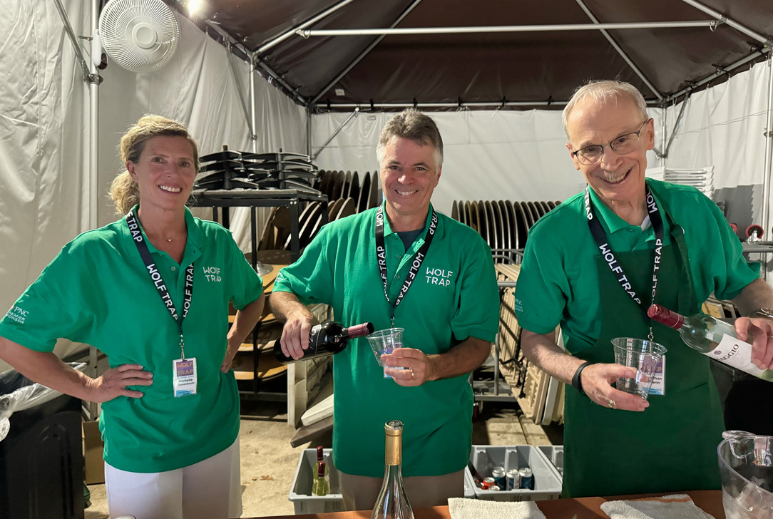 Three Wolf Trap volunteers in green shirts behind a table pouring wine inside a tent.