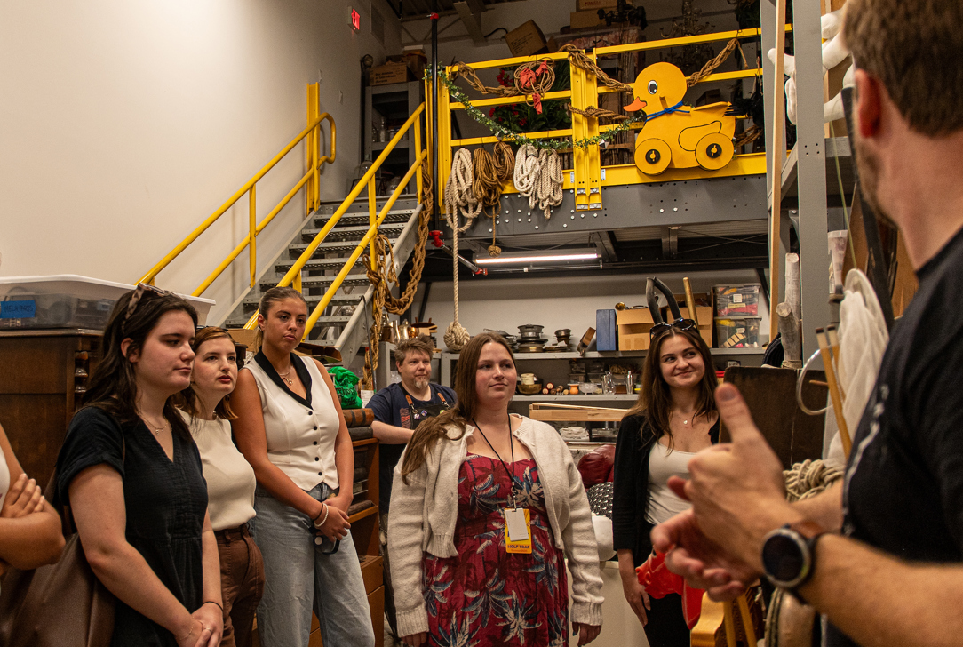 Interns gather in a prop storage area with shelves of ropes, tools, and stage equipment, while a staff member gestures during the tour.