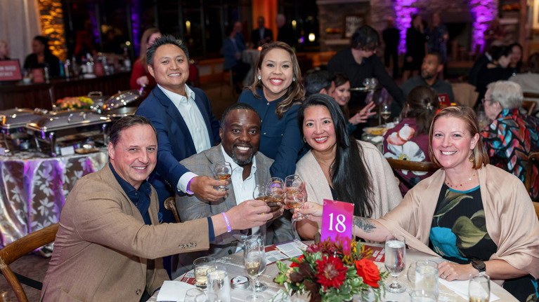 Group raising glasses in a toast around a decorated table with flowers and event signage.