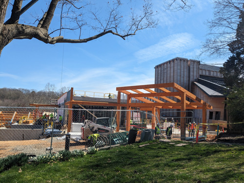 Wooden framework for an outdoor structure in progress, with workers and equipment behind a safety fence.